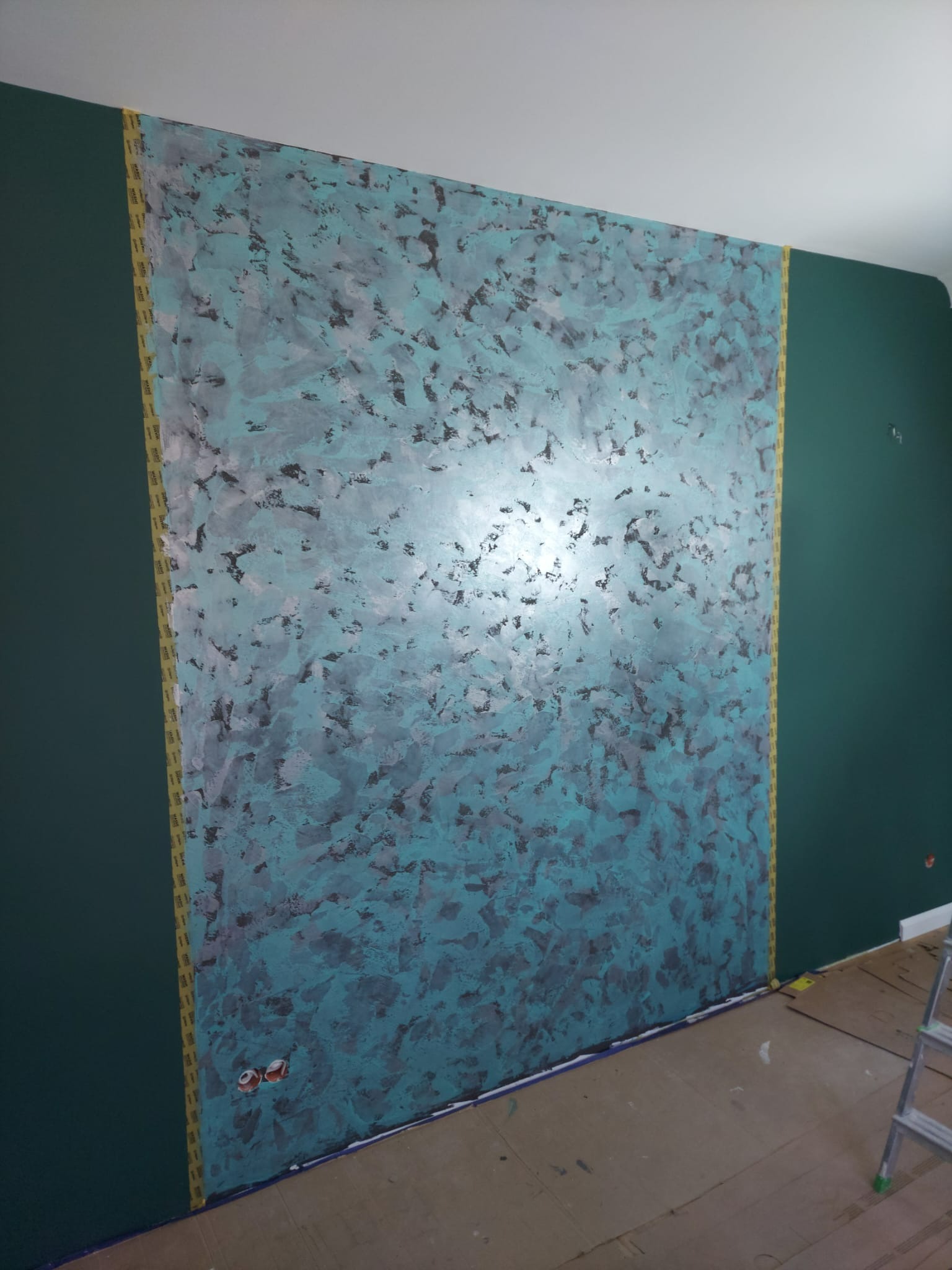 Ściana z dekoracyjną, turkusowo-szarą strukturą, zabezpieczona taśmą malarską. Obok ściana pomalowana na ciemnozielono. Widoczny fragment podłogi zabezpieczonej kartonem.