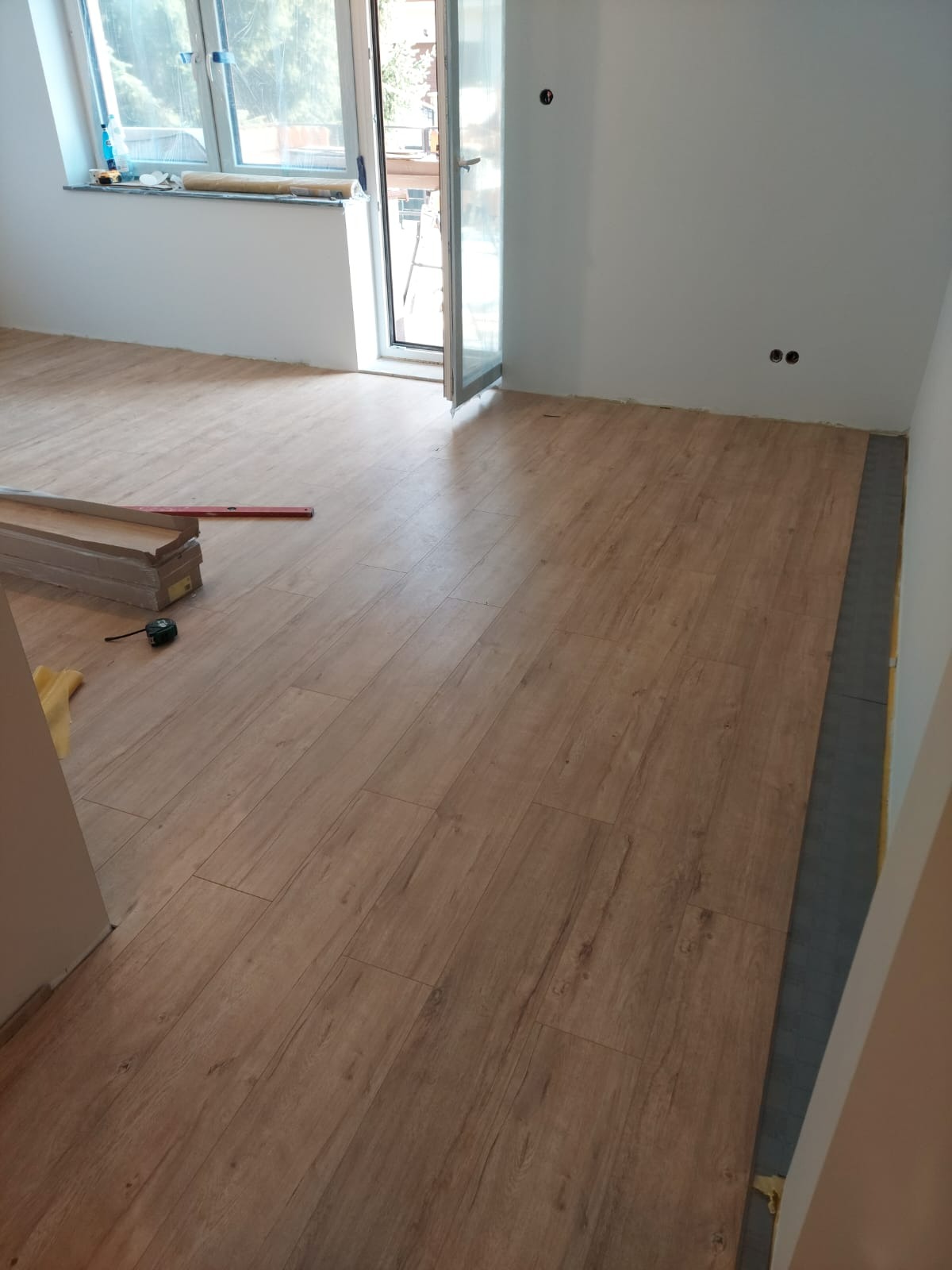 Układanie paneli podłogowych w jasnym wnętrzu. Widoczne panele, poziomica i miarka. Częściowo ułożona podłoga, okno i otwarte drzwi balkonowe.