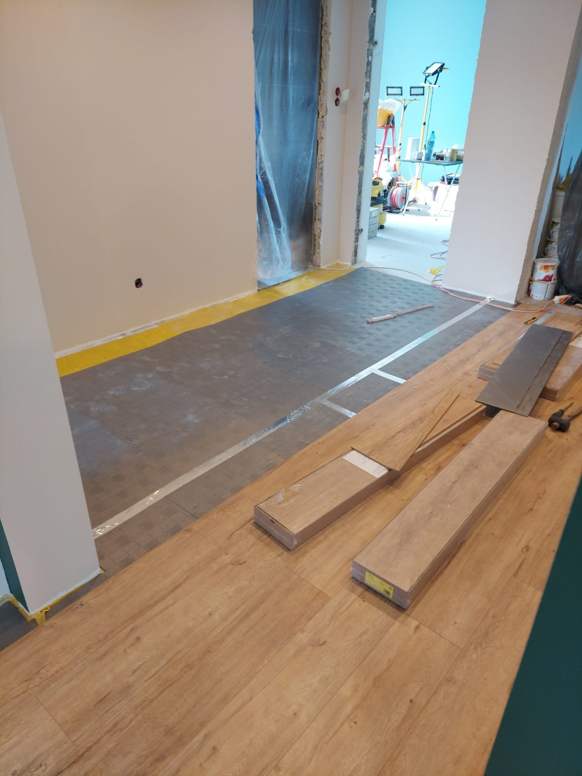 Układanie paneli podłogowych w trakcie remontu. Widoczna mata podkładowa i częściowo ułożone panele. W tle zabezpieczone folią drzwi i sprzęt remontowy.