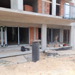 MILANOS PAWEŁ WYDRA - Montaż stolarki okiennej w surowym budynku. Widoczne profile okienne, narzędzia, pracownik w tle. Budowa w trakcie wykończenia.