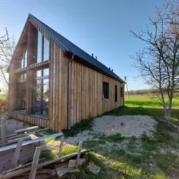 Nowoczesny dom z drewnianą elewacją i dużymi oknami, otoczony zielonym trawnikiem i elementami placu zabaw. Ujęcie w słoneczny dzień.