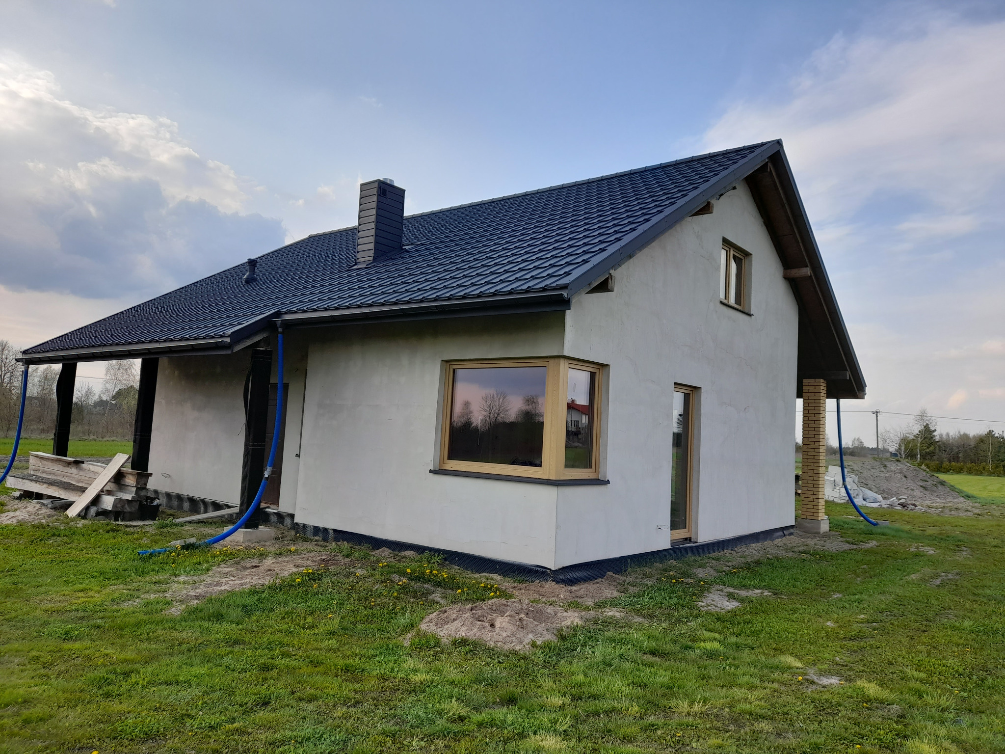 Dom jednorodzinny w trakcie wykończenia z drewnianą stolarką okienną, widoczne rury i materiały budowlane na zewnątrz. Ujęcie z boku, w tle pochmurne niebo.