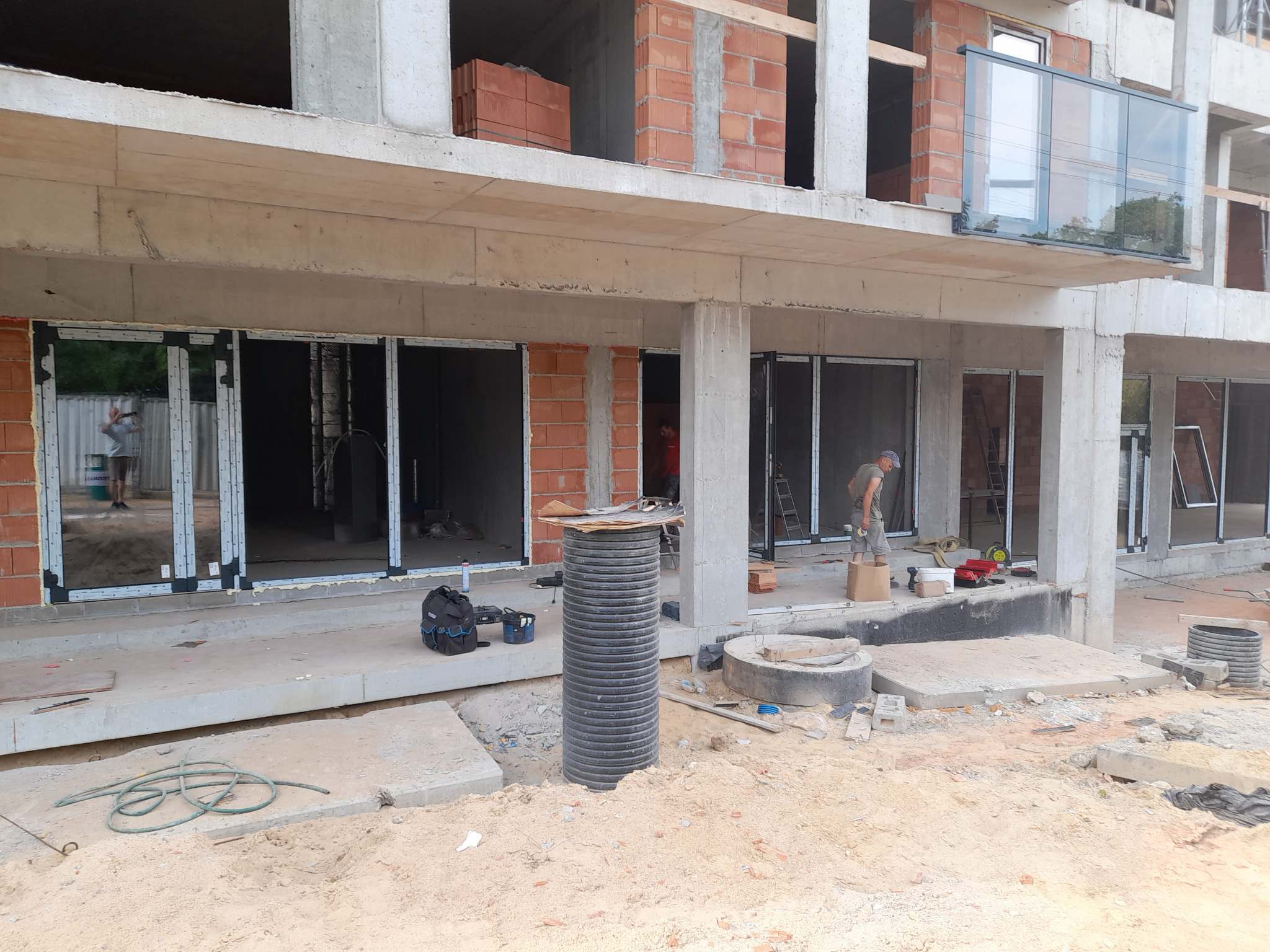 Montaż stolarki okiennej w surowym budynku. Widoczne profile okienne, narzędzia, pracownik w tle. Budowa w trakcie wykończenia.