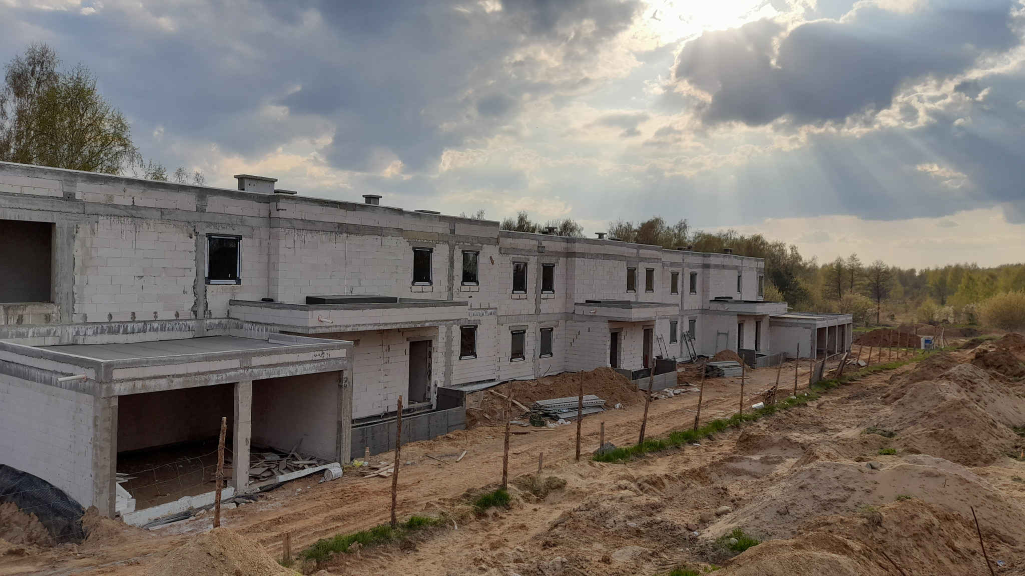 Surowy stan szeregowca z zamontowanymi oknami, widok z zewnątrz w trakcie budowy, teren niezagospodarowany, w tle drzewa i pochmurne niebo.