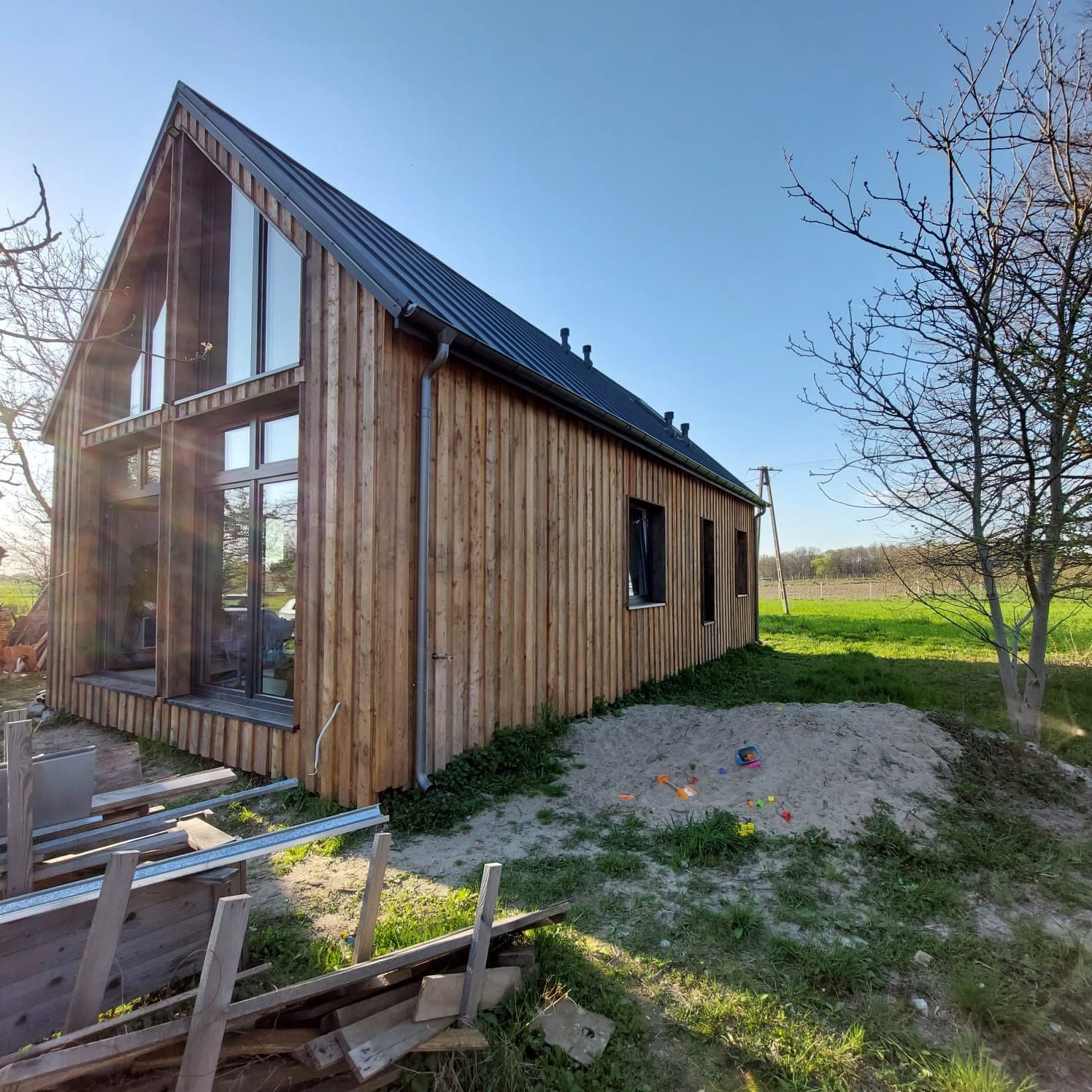 Nowoczesny dom z drewnianą elewacją i dużymi oknami, otoczony zielonym trawnikiem i elementami placu zabaw. Ujęcie w słoneczny dzień.