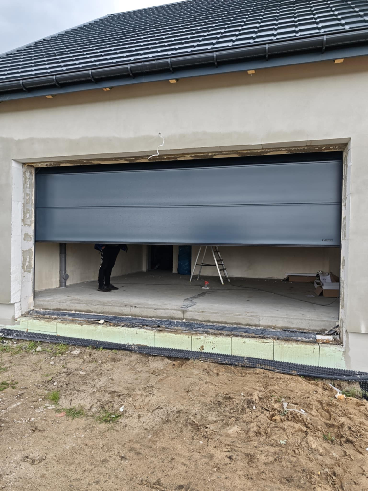 Montaż grafitowej bramy garażowej segmentowej w nowym budynku. Widoczne elementy konstrukcyjne i drabina w surowym garażu, z zewnątrz ziemia.