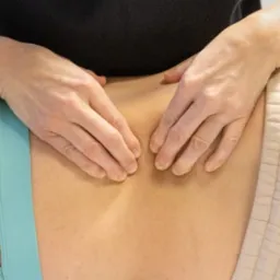 Dłonie terapeuty uciskają dolną część pleców pacjenta leżącego na beżowym podłożu, widoczny fragment turkusowej bielizny z ozdobnymi paskami.