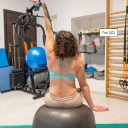 Fizjo TiM Łódź rehabilitacja - masaż - Kobieta siedzi na piłce rehabilitacyjnej w Łodzi, ćwicząc z gumą oporową, widok z tyłu na tle drabinek i sprzętu do ćwiczeń.