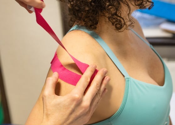 Aplikacja różowej taśmy kinesiotaping na ramieniu kobiety w błękitnym topie sportowym, widoczne kręcone włosy i dłoń terapeuty.
