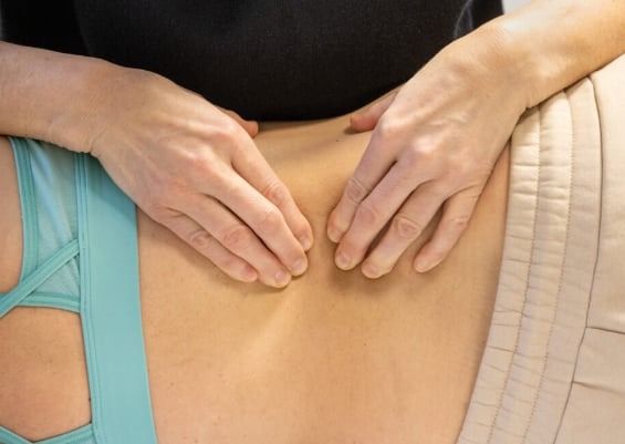 Dłonie terapeuty uciskają dolną część pleców pacjenta leżącego na beżowym podłożu, widoczny fragment turkusowej bielizny z ozdobnymi paskami.