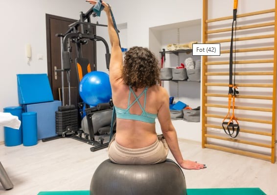 Kobieta siedzi na piłce rehabilitacyjnej w Łodzi, ćwicząc z gumą oporową, widok z tyłu na tle drabinek i sprzętu do ćwiczeń.
