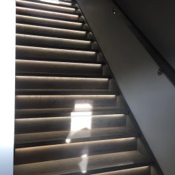 Wióry i opiłki - Nowoczesne schody z drewna oświetlone od spodu, z minimalistyczną metalową poręczą i geometrycznym obrazem na ścianie w tle.