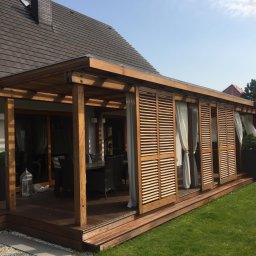 Wióry i opiłki - Drewniany taras z zadaszeniem i przesuwanymi żaluzjami oraz zasłonami, widok na stół z krzesłami i fragment domu z brązowym dachem.