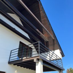 Wióry i opiłki - Nowoczesny balkon z czarną, metalową balustradą, wsparty na białych kolumnach, z drewnianym zadaszeniem i widokiem na jesienne drzewa.