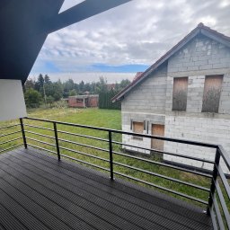 Wióry i opiłki - Czarny taras z desek kompozytowych i metalową balustradą, widok na budynek w stanie surowym i zarośniętą działkę.