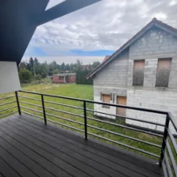 Czarny taras z desek kompozytowych i metalową balustradą, widok na budynek w stanie surowym i zarośniętą działkę.