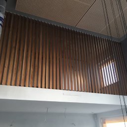 Wióry i opiłki - Montaż drewnianej ażurowej balustrady na antresoli z widocznym fragmentem okna i elementami konstrukcyjnymi zabezpieczonymi folią.