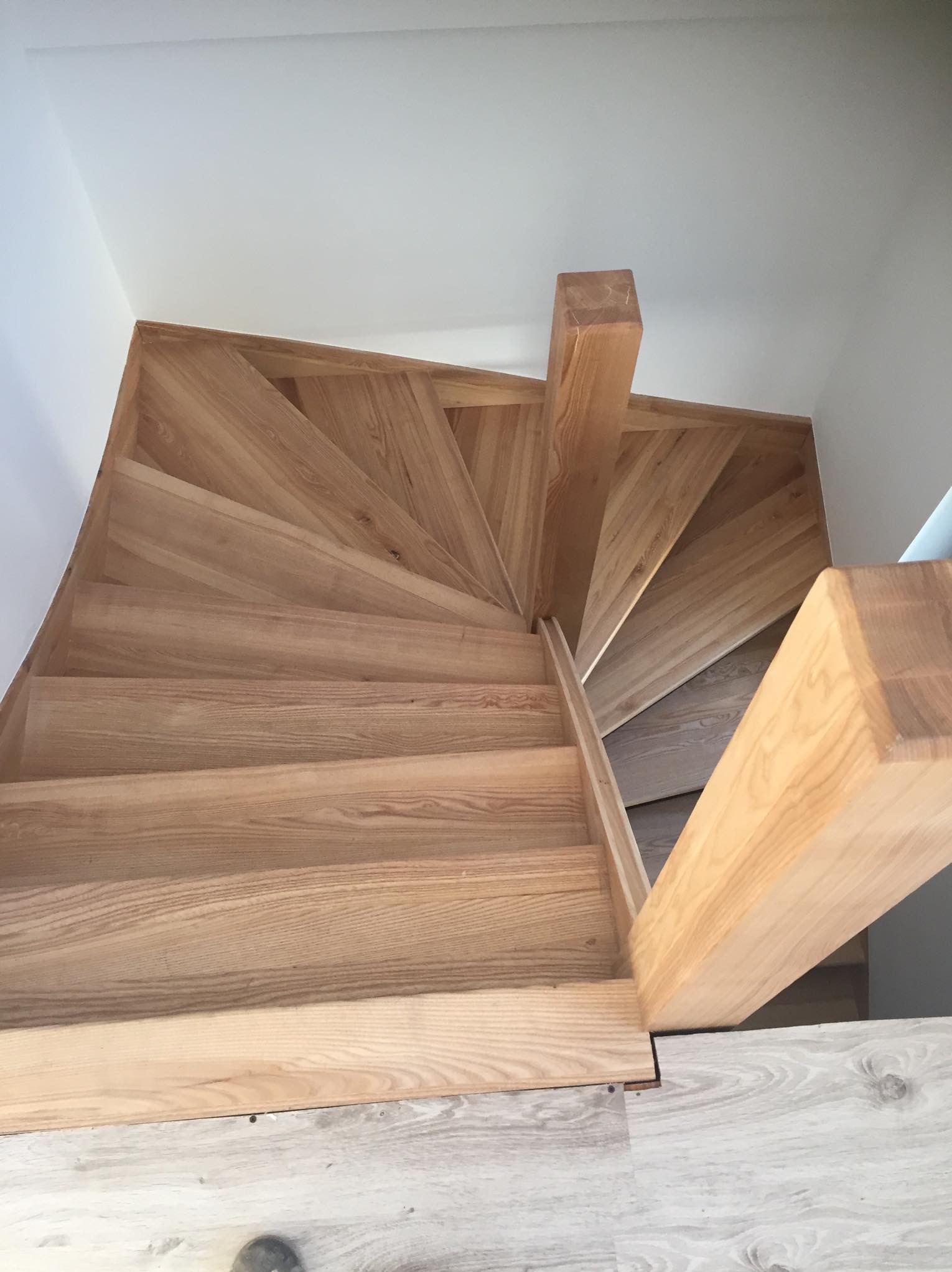 Spiralne schody z jasnego drewna, widok z góry, ukazujące stopnie i balustradę, z widocznym fragmentem podłogi wykończonej panelami imitującymi drewno.