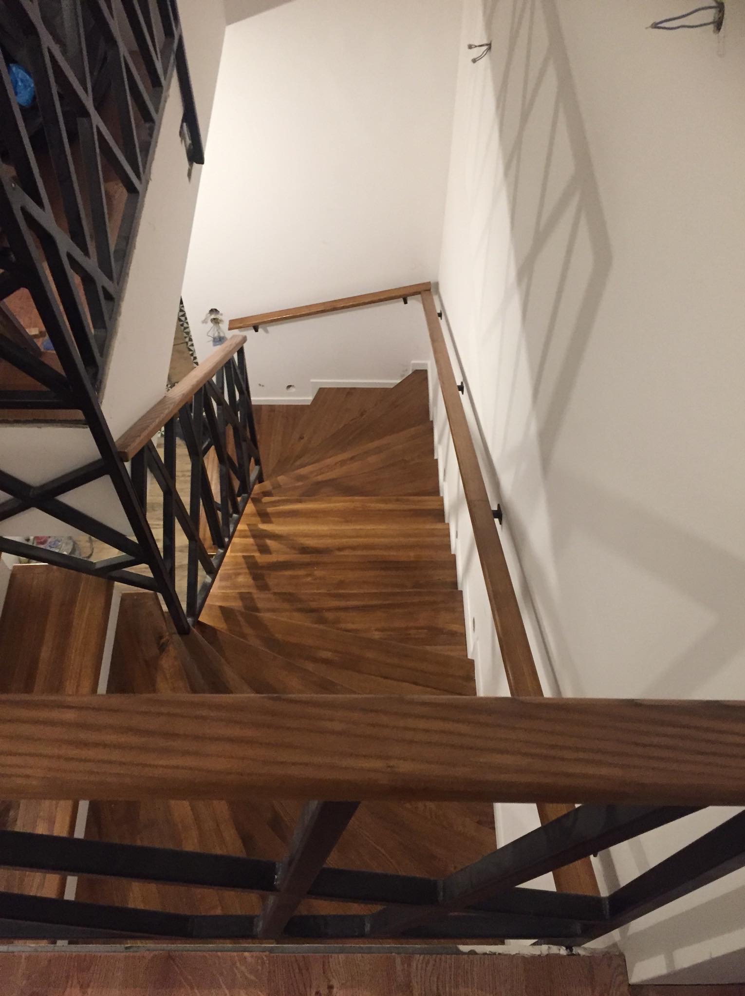 Wewnętrzne schody z drewna o spiralnym kształcie, z czarną metalową balustradą i drewnianą poręczą, widok z góry na dół, białe ściany, widoczne niedoskonałości wykończenia.