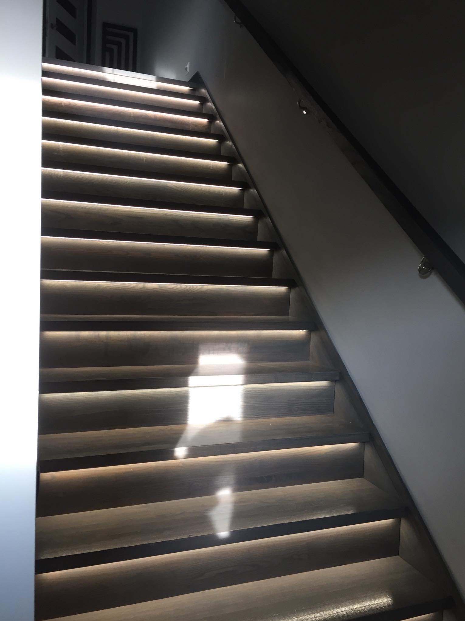 Nowoczesne schody z drewna oświetlone od spodu, z minimalistyczną metalową poręczą i geometrycznym obrazem na ścianie w tle.