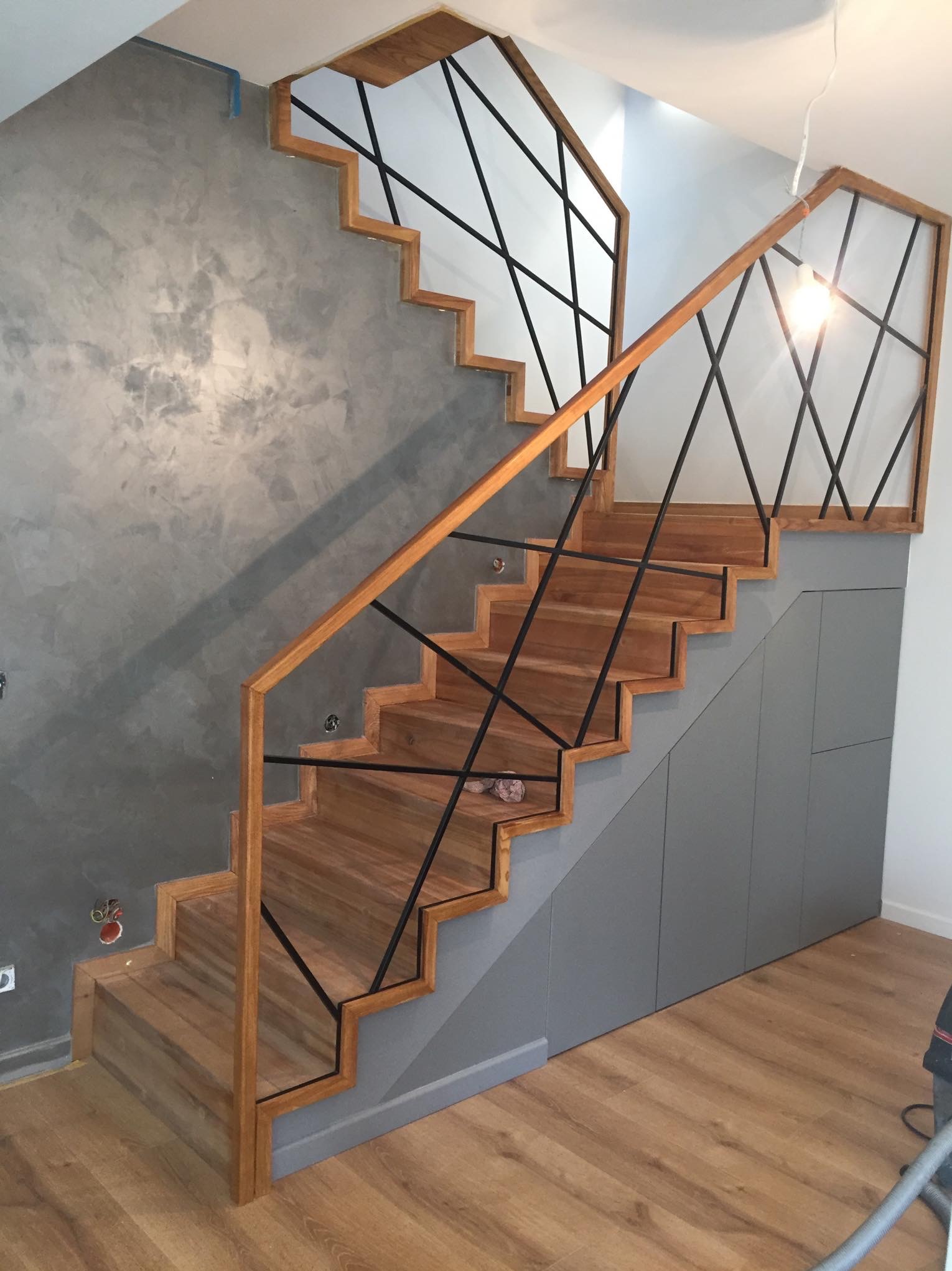 Nowoczesne schody drewniane z czarną, geometryczną balustradą na tle ściany w odcieniach szarości, zintegrowane z szarą zabudową szafkową pod biegiem schodów.