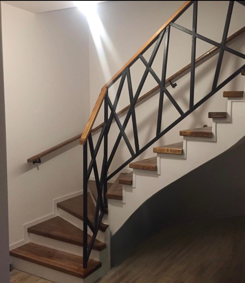 Wewnętrzne schody z drewnianymi stopniami i czarną, geometryczną balustradą ze szczebli, oświetlone z góry, z drewnianą poręczą.