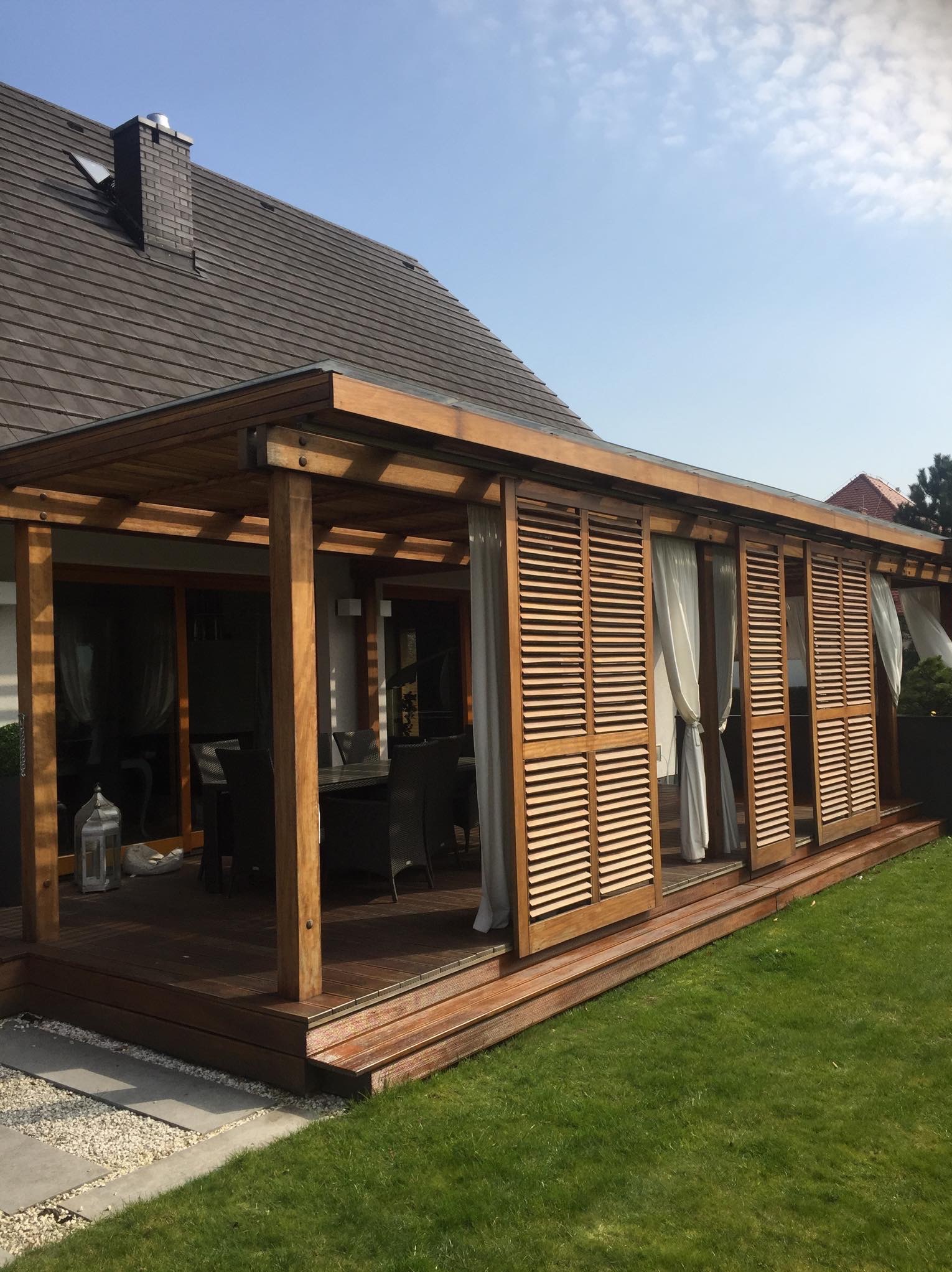 Drewniany taras z zadaszeniem i przesuwanymi żaluzjami oraz zasłonami, widok na stół z krzesłami i fragment domu z brązowym dachem.