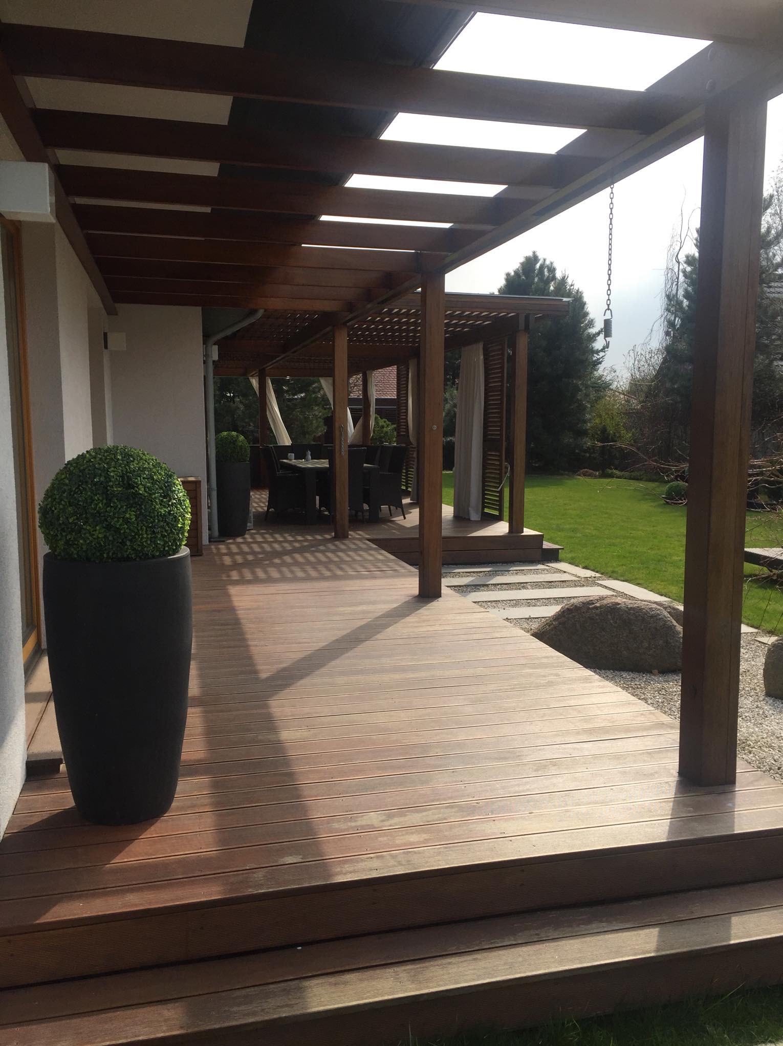 Zadaszony taras z drewnianą podłogą i pergolą, oświetlony promieniami słońca, z dekoracyjną kulą z zieleni w czarnej donicy i widokiem na zadbany ogród.