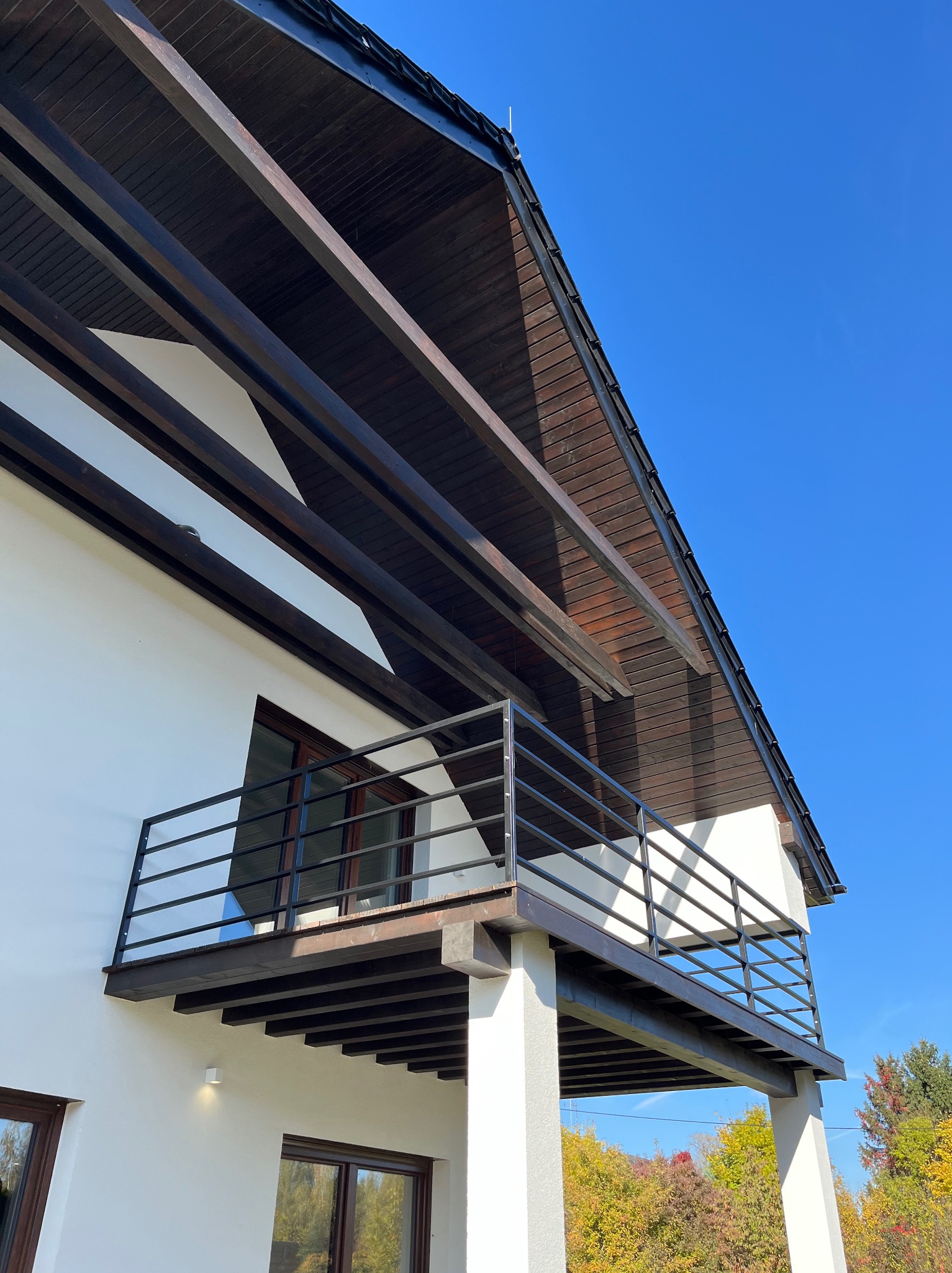 Nowoczesny balkon z czarną, metalową balustradą, wsparty na białych kolumnach, z drewnianym zadaszeniem i widokiem na jesienne drzewa.
