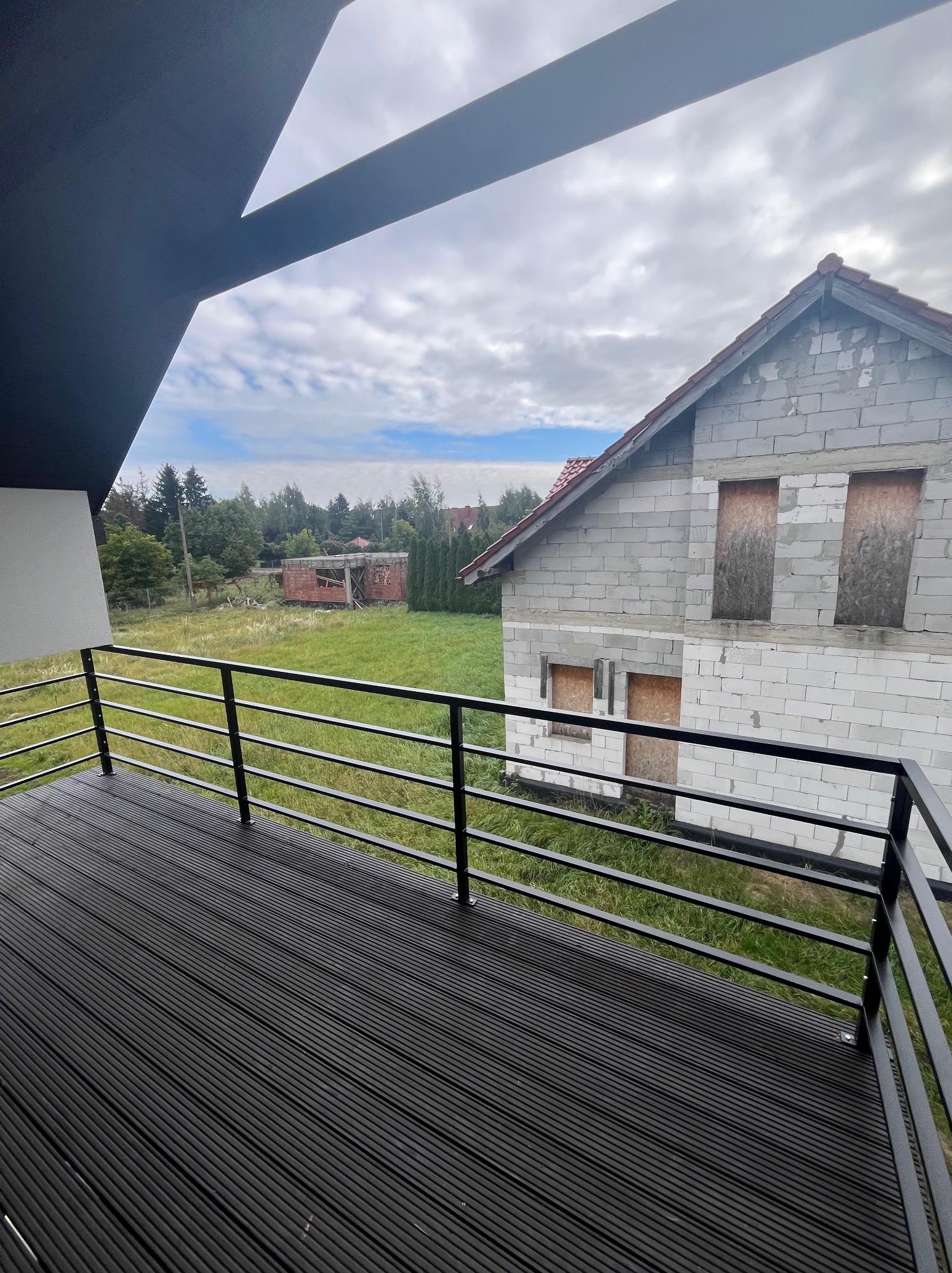 Czarny taras z desek kompozytowych i metalową balustradą, widok na budynek w stanie surowym i zarośniętą działkę.