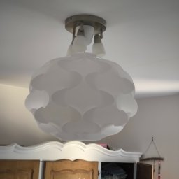 Jakub Jędrzejewski - Zamontowana biała lampa sufitowa o dekoracyjnym, ażurowym kloszu, widoczna na tle białego sufitu i fragmentu szafy z otwartymi drzwiami.