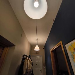 Jakub Jędrzejewski - Korytarz z dwiema lampami wiszącymi: duża okrągła i mała stożkowa, zamontowanymi natynkowo na suficie, ściany pomalowane na beżowo i granatowo, widoczny wieszak na ubrania i drzwi wejściowe.
