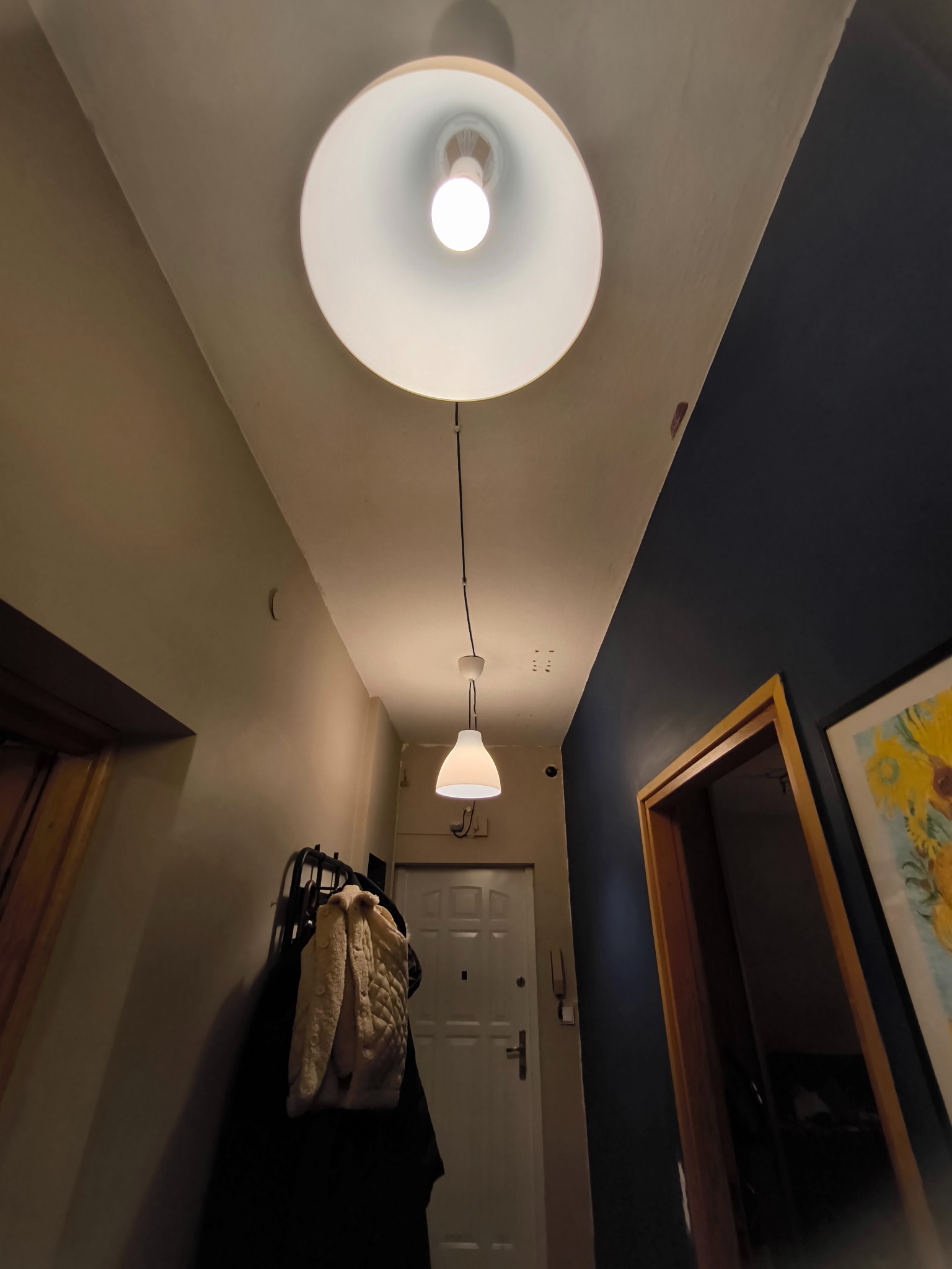 Korytarz z dwiema lampami wiszącymi: duża okrągła i mała stożkowa, zamontowanymi natynkowo na suficie, ściany pomalowane na beżowo i granatowo, widoczny wieszak na ubrania i drzwi wejściowe.