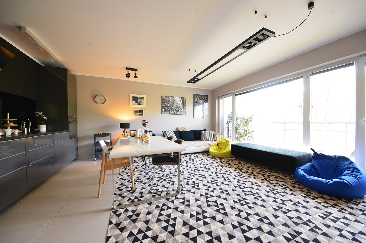 Nowoczesne oświetlenie sufitowe w przestronnym salonie z aneksem kuchennym w Poznaniu. Widoczny stół z krzesłami, sofa, pufy i wzorzysty dywan.