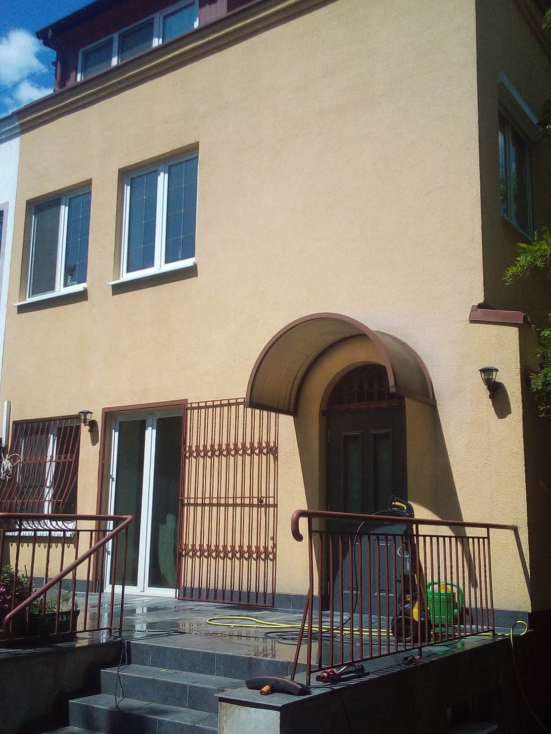 Dwupiętrowy dom z beżową elewacją, widoczna myjka ciśnieniowa na schodach wejściowych, metalowa krata przy drzwiach balkonowych, oraz mokry, ciemny bruk na schodach i przed wejściem.