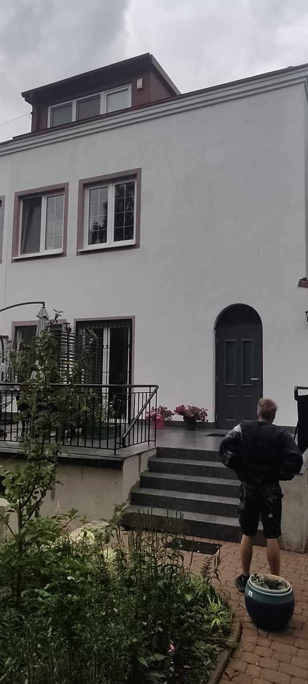 Dwupiętrowy dom z białymi ścianami i szarą stolarką okienną, widok z poziomu ogrodu, osoba w ubraniu roboczym stoi obok donicy.