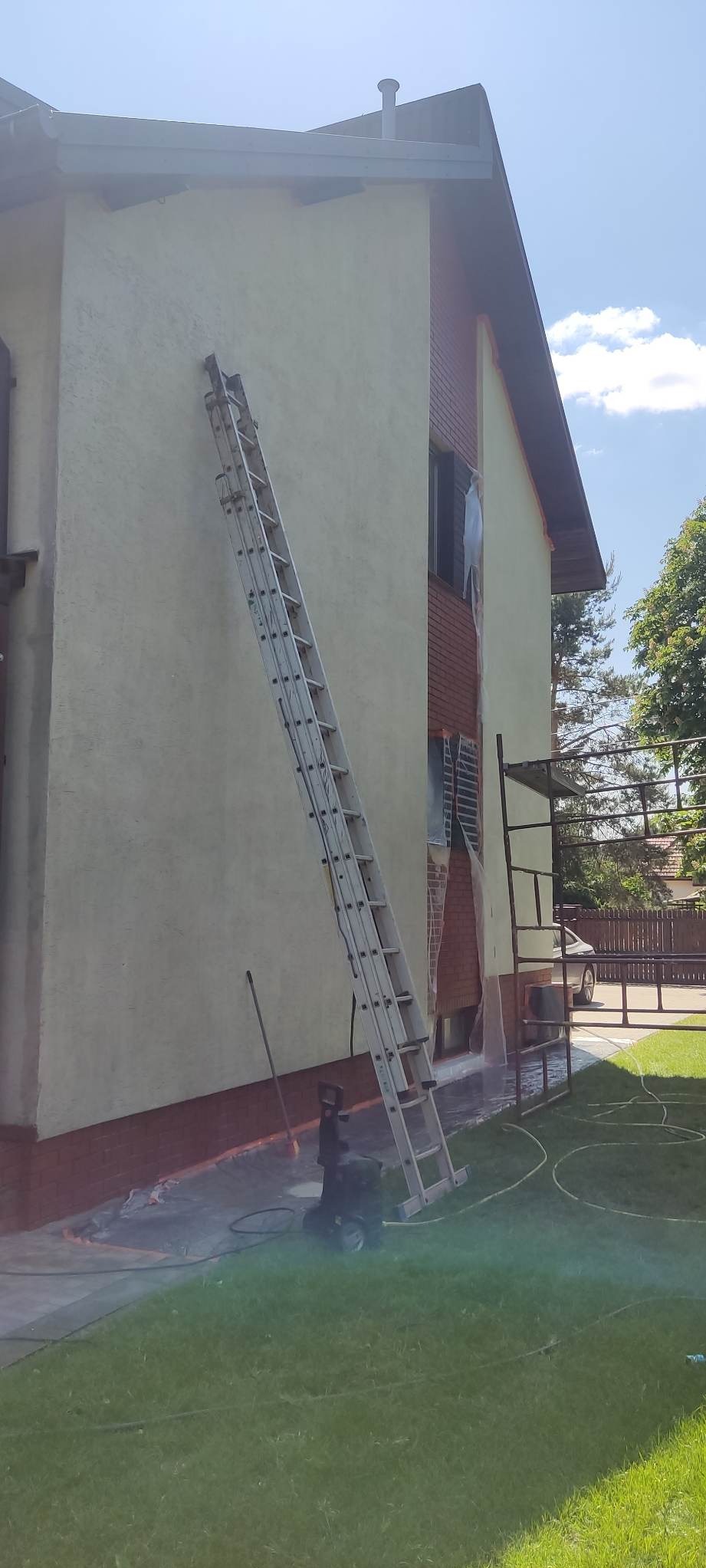 Mycie elewacji domu jednorodzinnego z użyciem myjki ciśnieniowej, widoczna drabina oparta o ścianę, okna zabezpieczone folią.