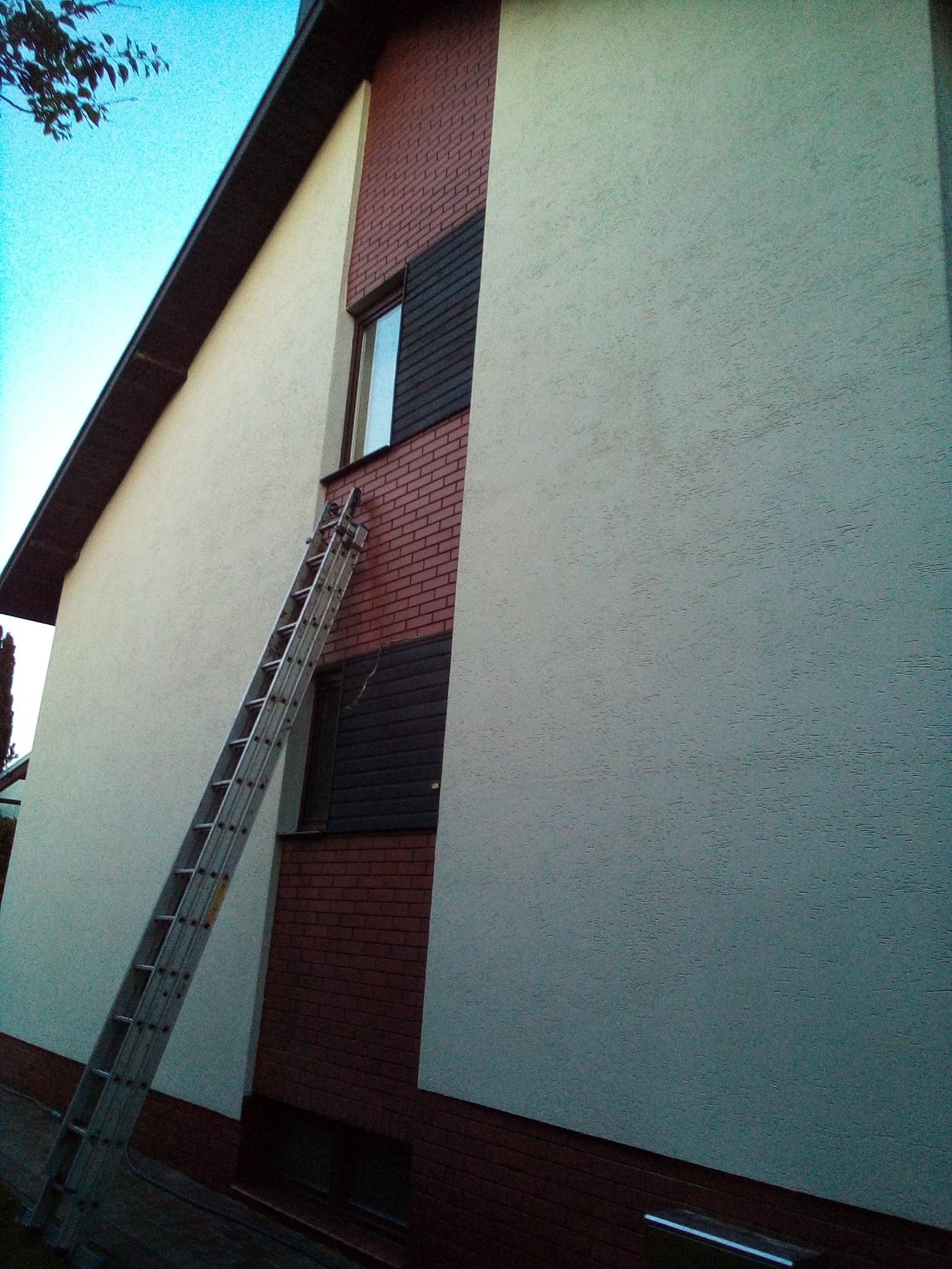 Dwupiętrowy dom z elewacją w odcieniach bieli i ceglanej czerwieni, z przystawioną aluminiową drabiną do okna na piętrze, widok z dołu.