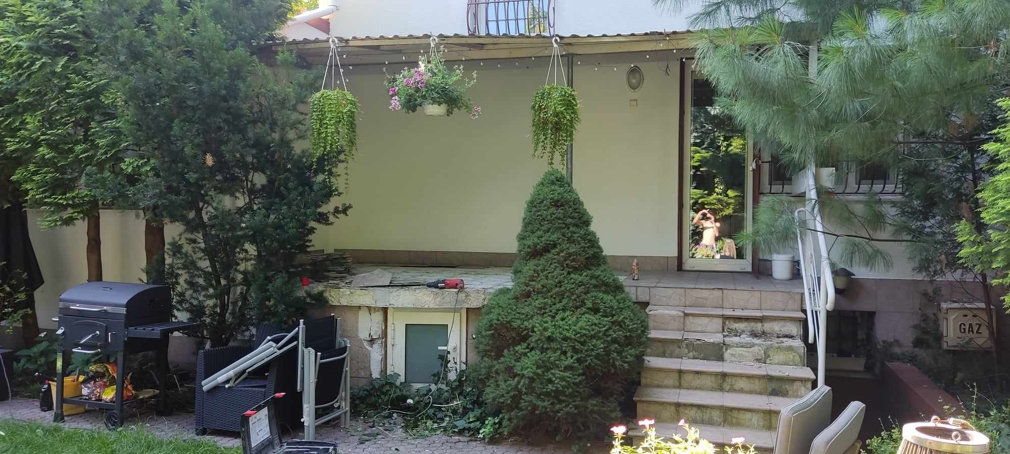 Przygotowania do remontu elewacji budynku jednorodzinnego: narzędzia, płyty elewacyjne, grill i meble ogrodowe zgromadzone przed wejściem.