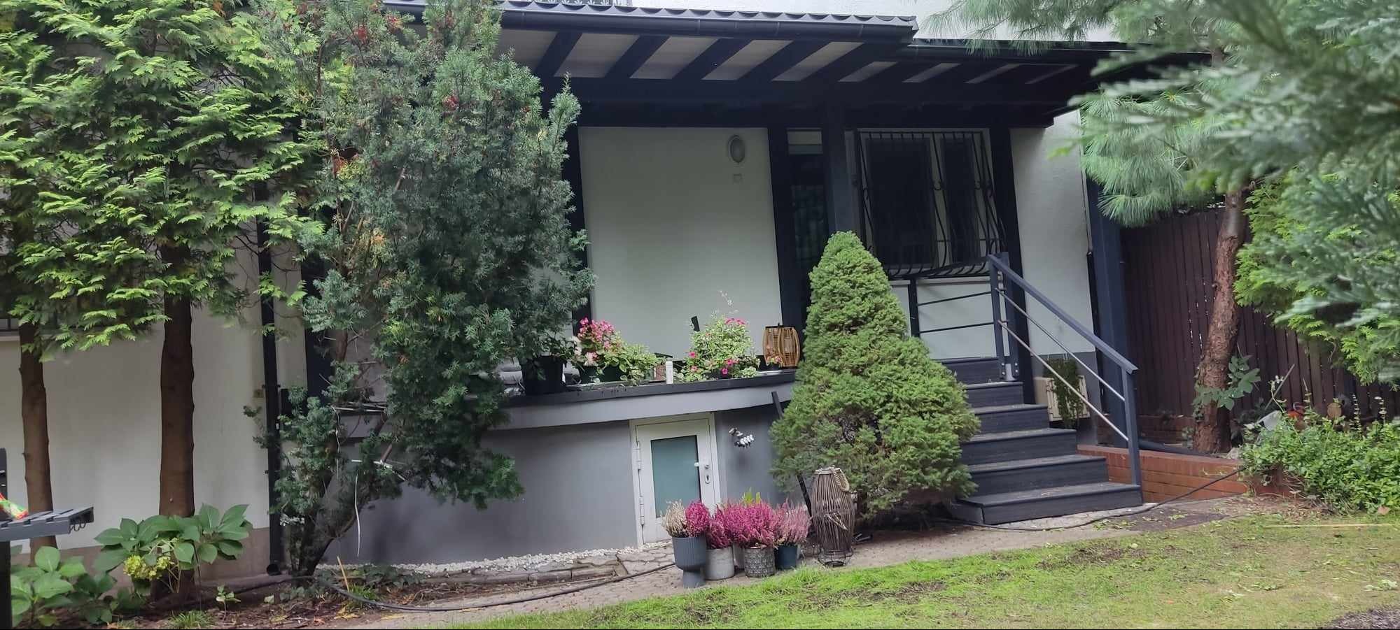 Jasnoszary dom z ciemnym dachem i schodami, ozdobiony roślinami w donicach i drzewami iglastymi.