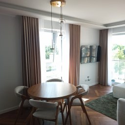 NAVA Vasyl Nakoniushevskyi - Nowoczesny salon z jadalnią: okrągły drewniany stół z czterema tapicerowanymi krzesłami, parkiet w jodełkę, designerska lampa wisząca i beżowe zasłony.