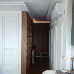 NAVA Vasyl Nakoniushevskyi - Jasne wnętrze z białymi ścianami ze sztukaterią, drewnianą podłogą w jodełkę i wbudowanym regałem. Sufit z ukrytym oświetleniem LED i klimatyzatorem ściennym.