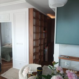 NAVA Vasyl Nakoniushevskyi - Eleganckie wnętrze z białymi ścianami ze sztukaterią, wbudowanym drewnianym regałem i bukietem różowych piwonii na stole. W tle widoczna sypialnia z zielonym łóżkiem.