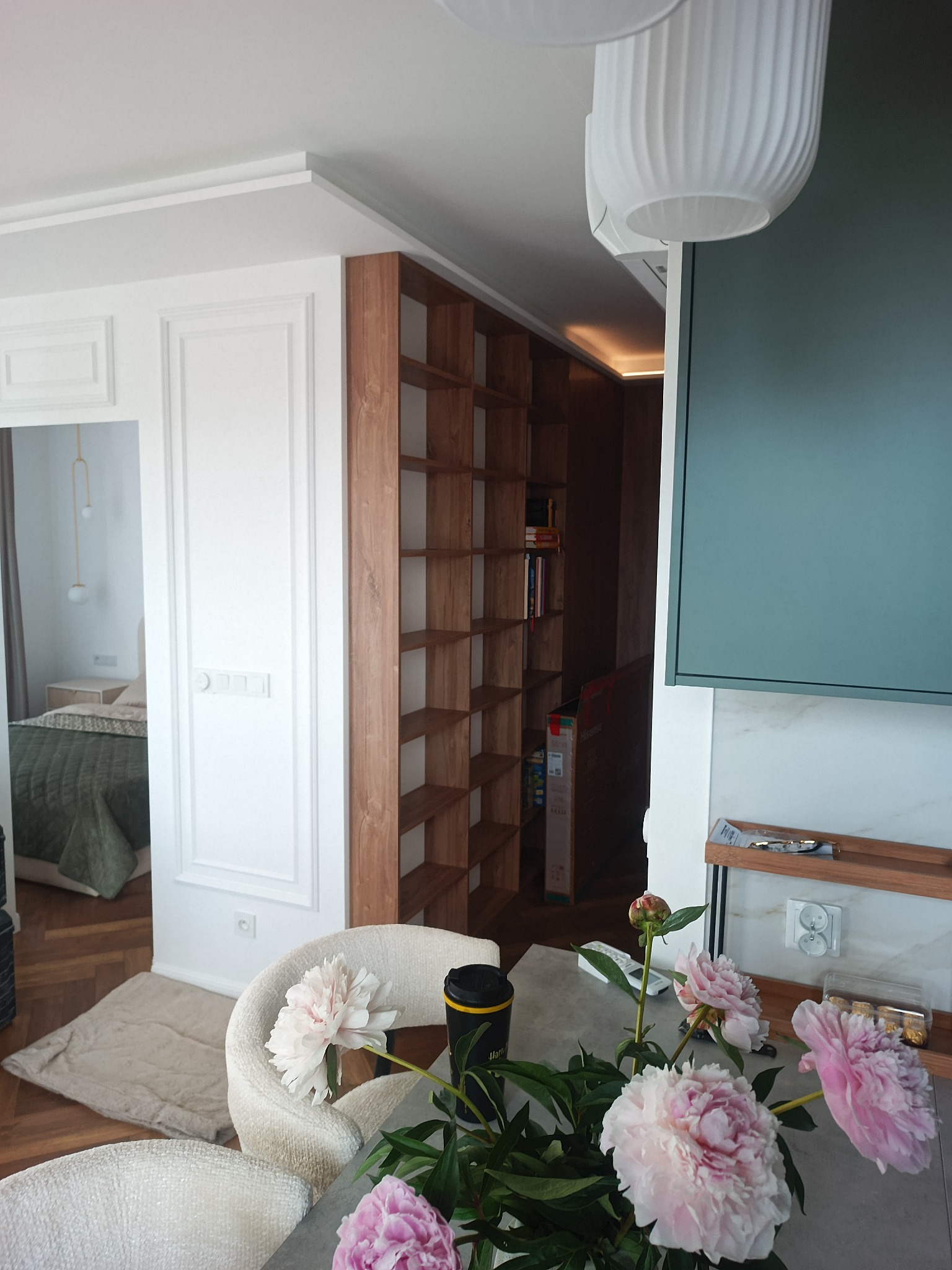 Eleganckie wnętrze z białymi ścianami ze sztukaterią, wbudowanym drewnianym regałem i bukietem różowych piwonii na stole. W tle widoczna sypialnia z zielonym łóżkiem.