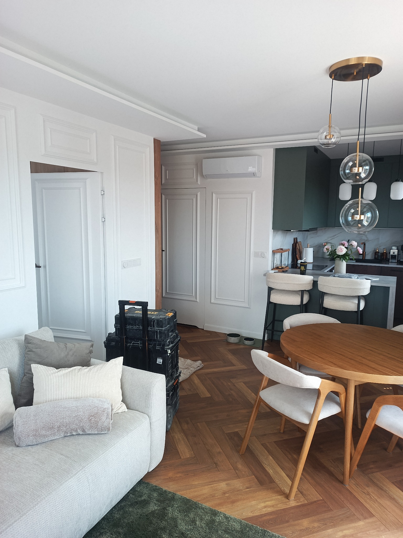 Nowoczesny salon z aneksem kuchennym: ciemnozielone szafki, złote lampy wiszące, drewniany stół, jasna sofa, białe panele ścienne i parkiet w jodełkę.