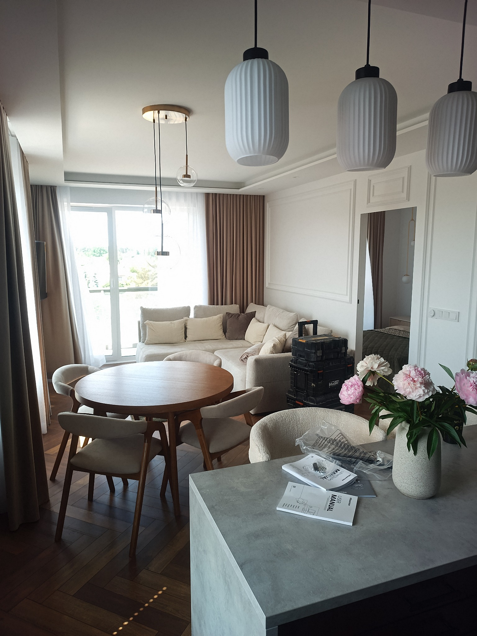 Nowoczesny salon z jadalnią, beżowa sofa, okrągły drewniany stół, podłoga w jodełkę. Sufit z oświetleniem, ściany ze sztukaterią. Widoczne narzędzia i instrukcje montażowe.