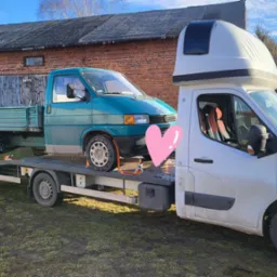 Zielony Volkswagen Transporter T4 na platformie lawety z białą kabiną i nadbudówką, na tle ceglanego budynku gospodarczego.