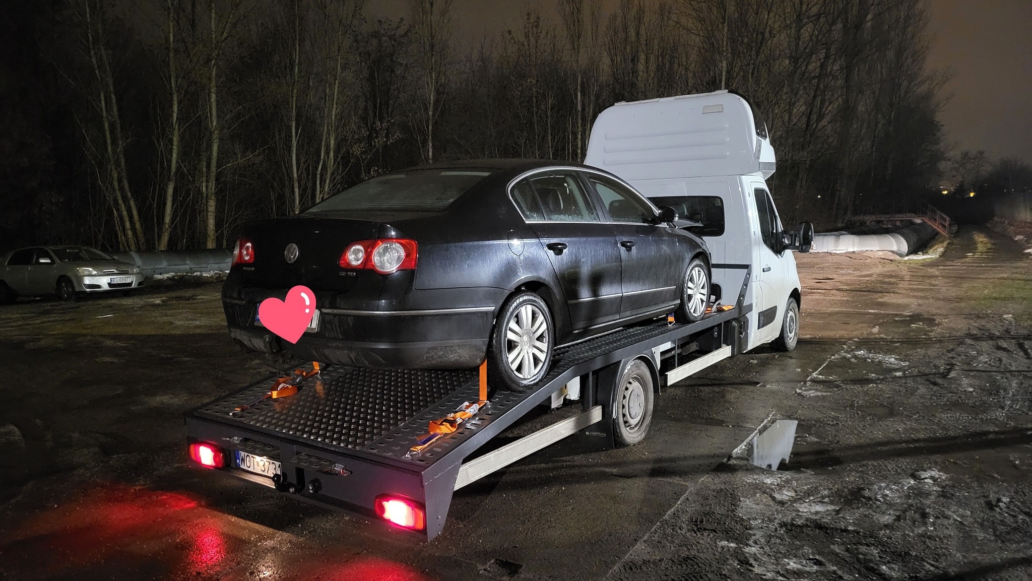 Czarny samochód osobowy marki Volkswagen Passat 2.0 TDI zabezpieczony pasami transportowymi na platformie lawety w warunkach nocnych; w tle widoczne drzewa i inny pojazd.