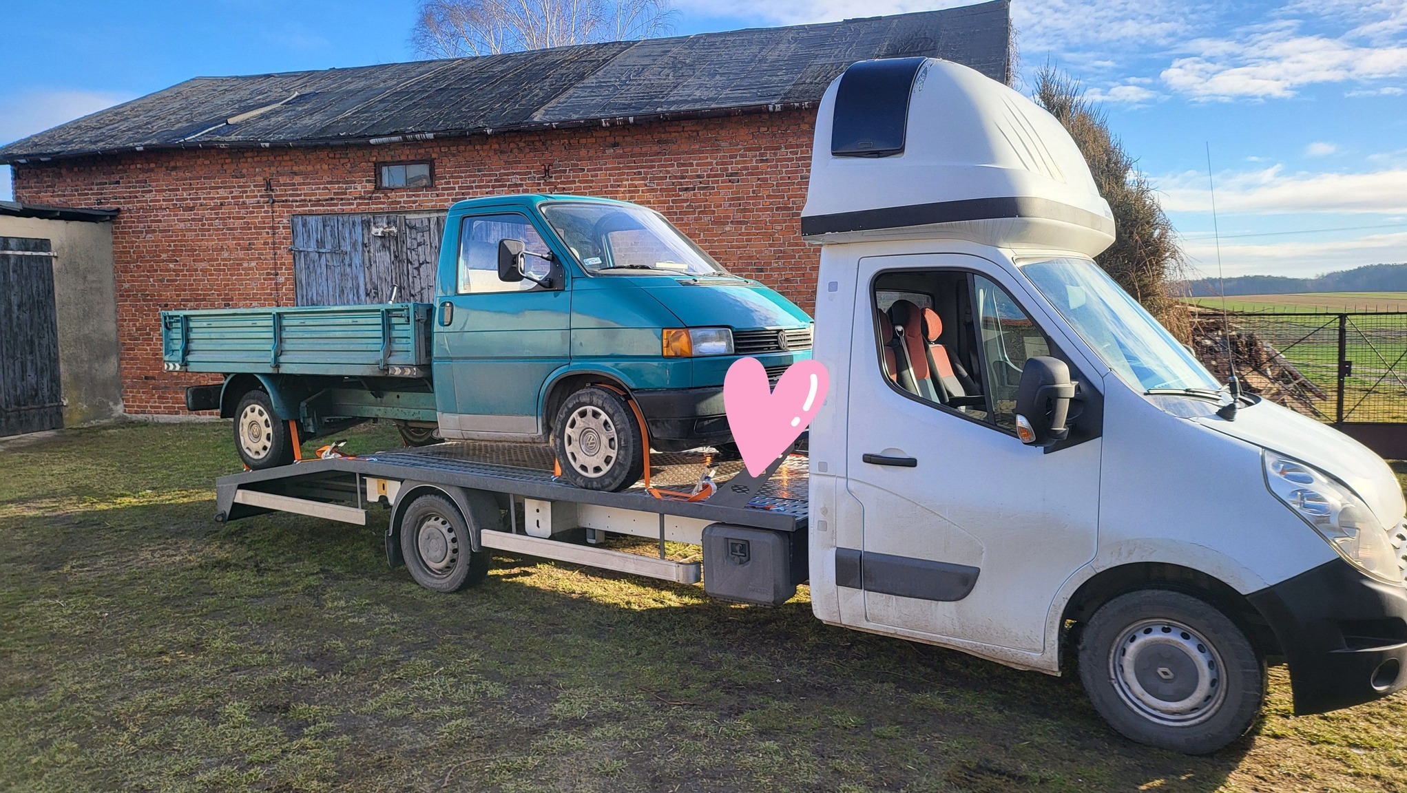 Zielony Volkswagen Transporter T4 na platformie lawety z białą kabiną i nadbudówką, na tle ceglanego budynku gospodarczego.