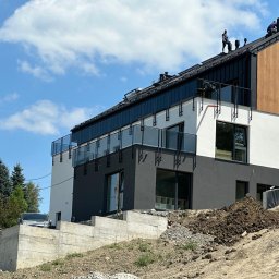 Uslugi ogolnobudowlane - Dwóch pracowników na dachu nowoczesnego domu z drewnianą elewacją, widok z dołu na tle błękitnego nieba z chmurami, teren przed domem w trakcie niwelacji.